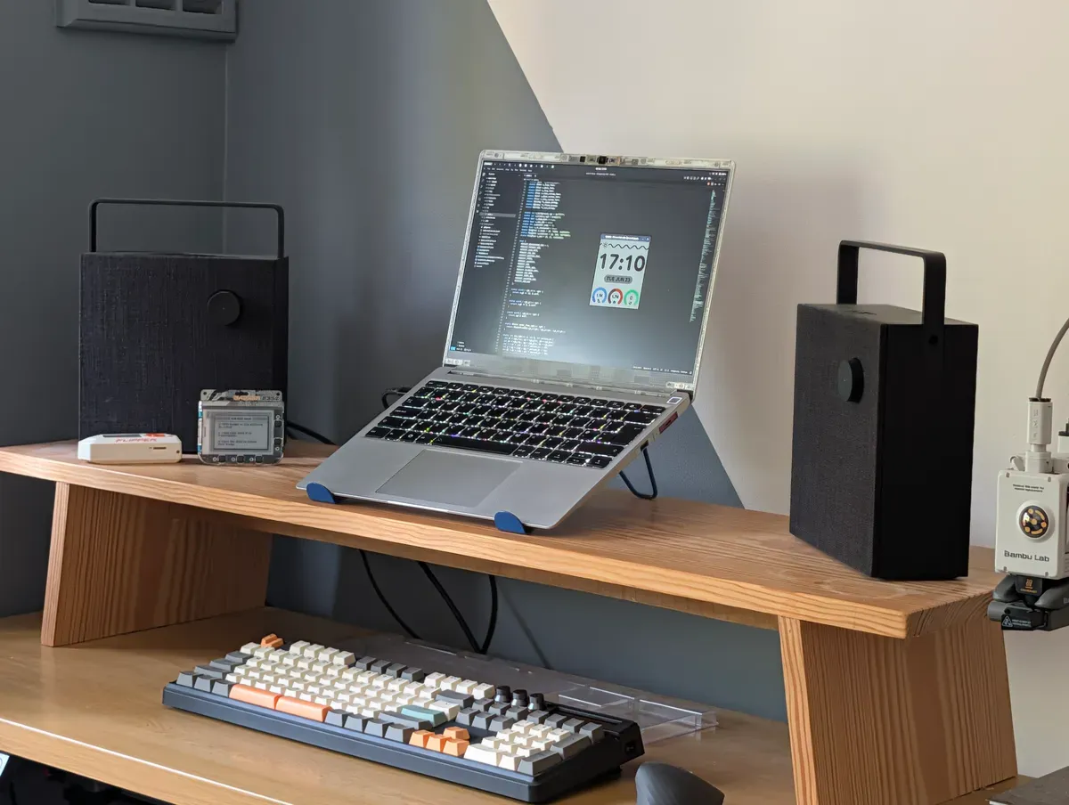 A desk setup featuring a Framework laptop on a wooden stand displaying a Pebble watchface emulator and code editor, flanked by two black speakers and a mechanical keyboard